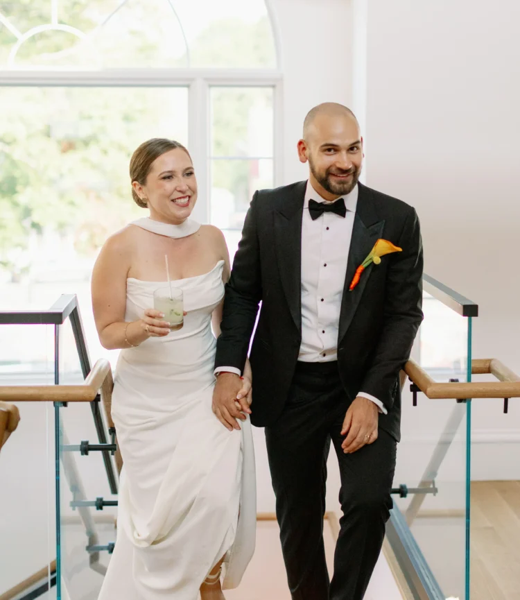 Couple celebrating at Belvedere Lenox, bride in elegant white gown holding cocktail, groom in tuxedo with boutonniere, joyful moment during wedding event.