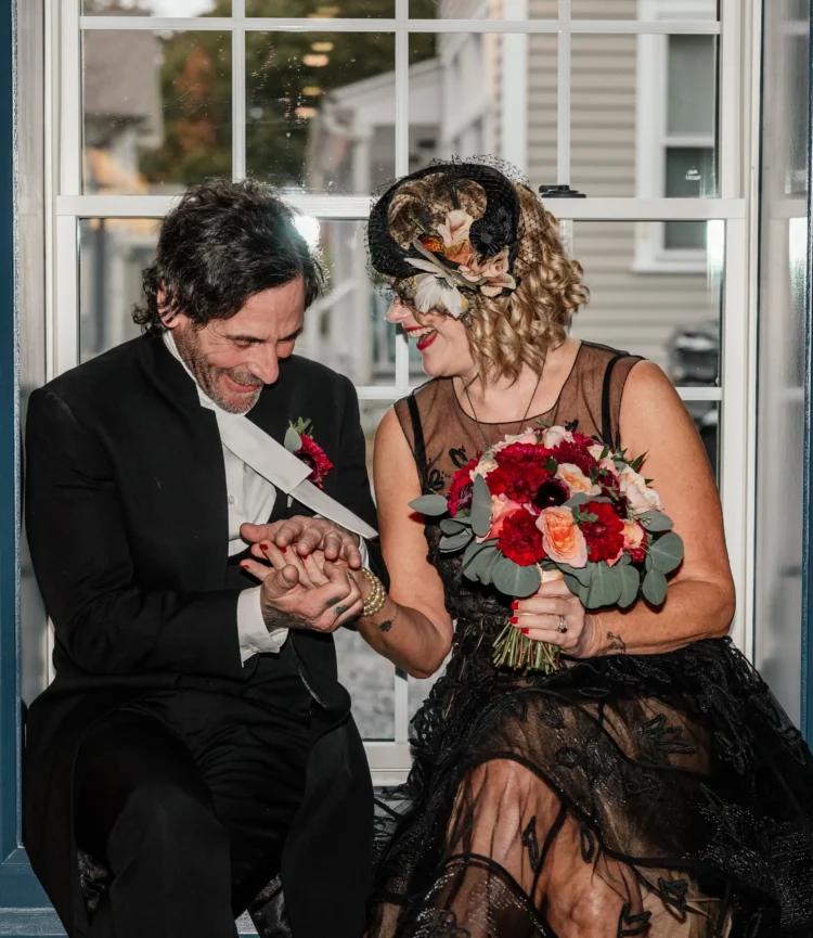Couple John and Kate sharing a joyful moment, holding hands, dressed in elegant wedding attire, with a bouquet of flowers, inside Belvedere Lenox venue.