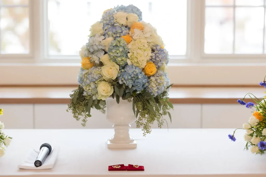 Floral arrangement featuring blue hydrangeas, white roses, and yellow accents in a decorative vase, with a microphone and decorative elements on a table, showcasing event decor at Belvedere Lenox venue.