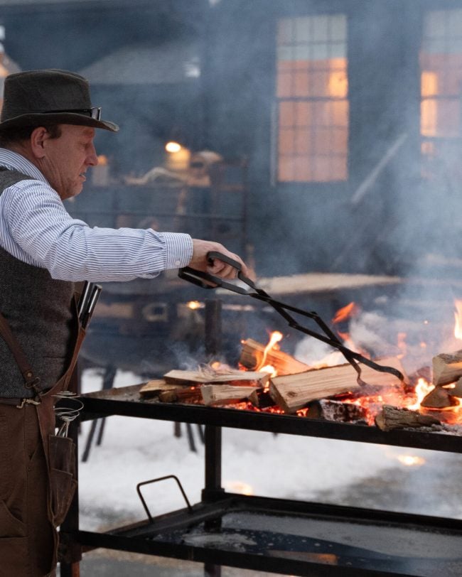 Man tending a wood fire outdoors, wearing a hat and apron, with flames and logs visible, creating a warm, inviting atmosphere for gatherings at Belvedere Lenox.