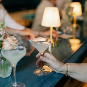 Cocktail glasses on a stylish bar at Belvedere Lenox, featuring a hand holding a drink, with soft lighting and floral arrangements, ideal for wedding rehearsal dinners and welcome events.