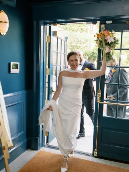 Bride in elegant white gown joyfully entering venue with bouquet, celebrating wedding moments at Belvedere Lenox, a sophisticated event space in the Berkshires.