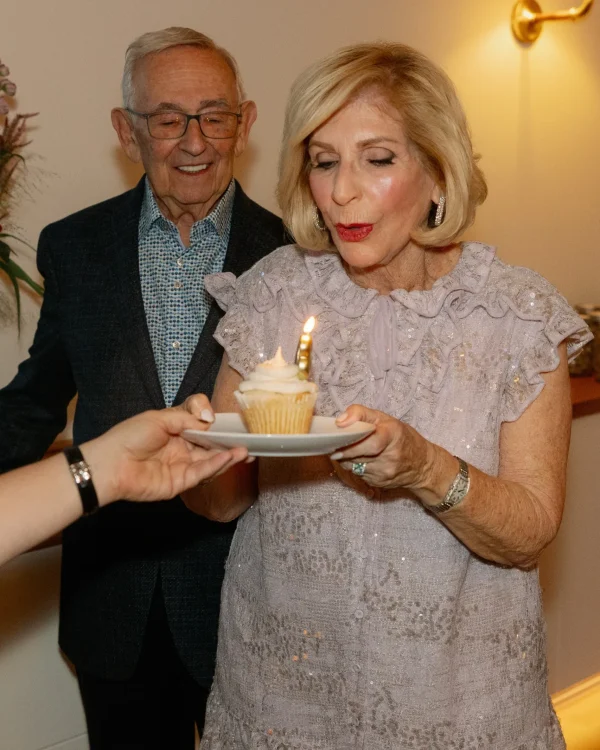Woman blowing out candle on cupcake, celebrating a birthday, with a man in the background, in a festive event setting.