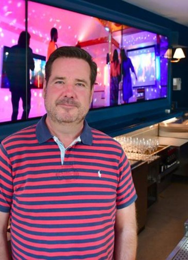 Man in a striped polo shirt standing in front of a vibrant digital screen displaying a lively event atmosphere, showcasing Belvedere Lenox as a premier venue for corporate gatherings and special occasions.