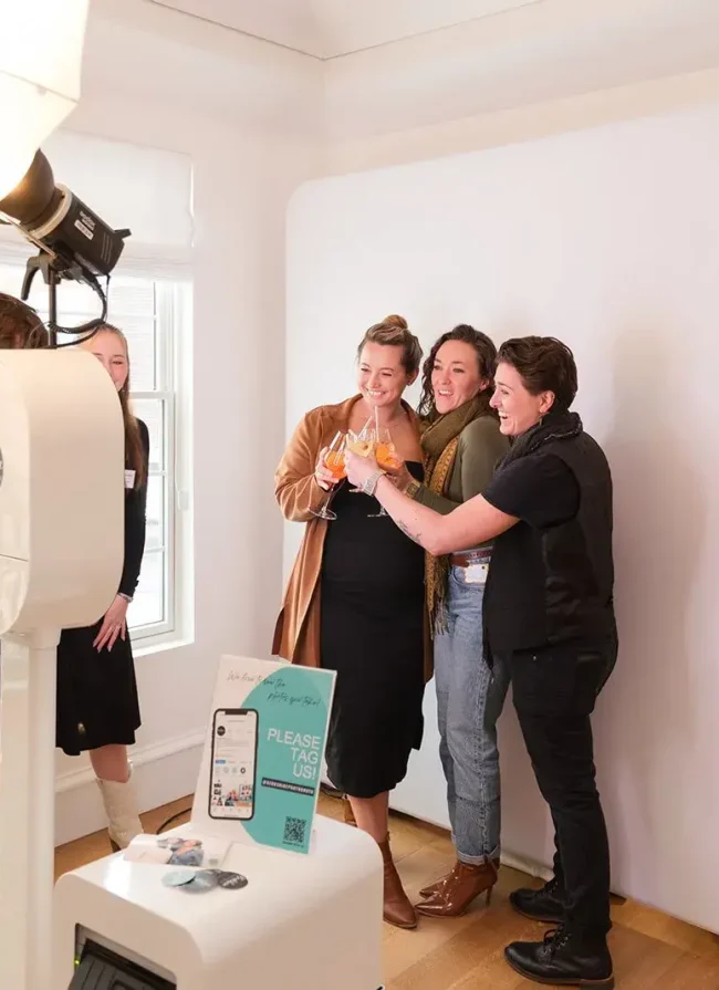Friends celebrating with cocktails in a photo booth at an event, featuring a sign encouraging guests to tag the venue on social media, highlighting the fun atmosphere of gatherings at Belvedere Lenox.