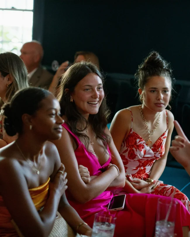 Three women enjoying a lively moment at an event, dressed in vibrant dresses, seated together with drinks on the table, capturing the essence of celebration and connection at Belvedere Lenox.